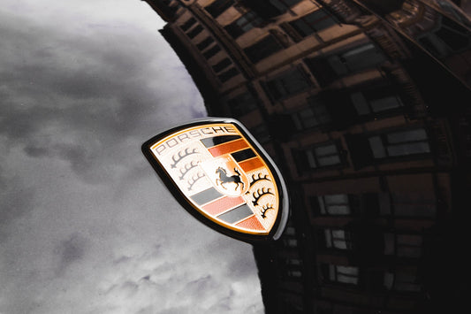 Porsche logo on black car hood with building and sky reflections, luxury sports car detail