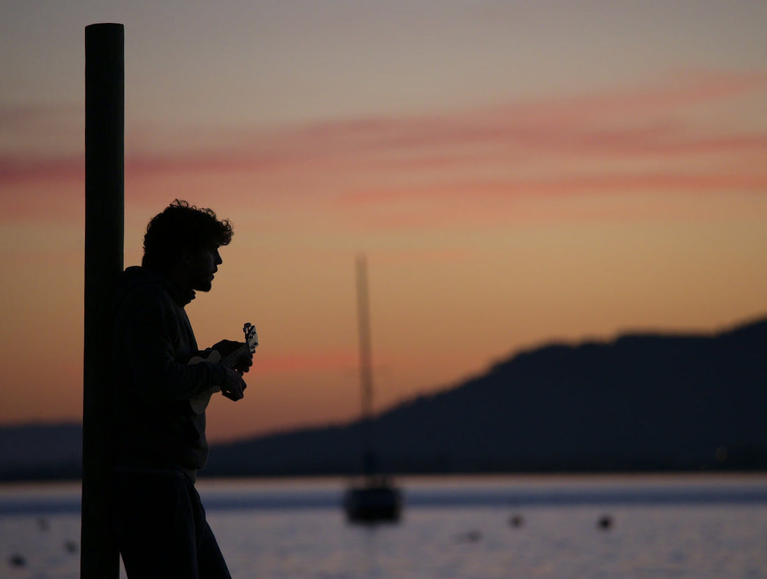 Silhouette of man playing ukulele by lake at sunset with mountains and sailboat in background