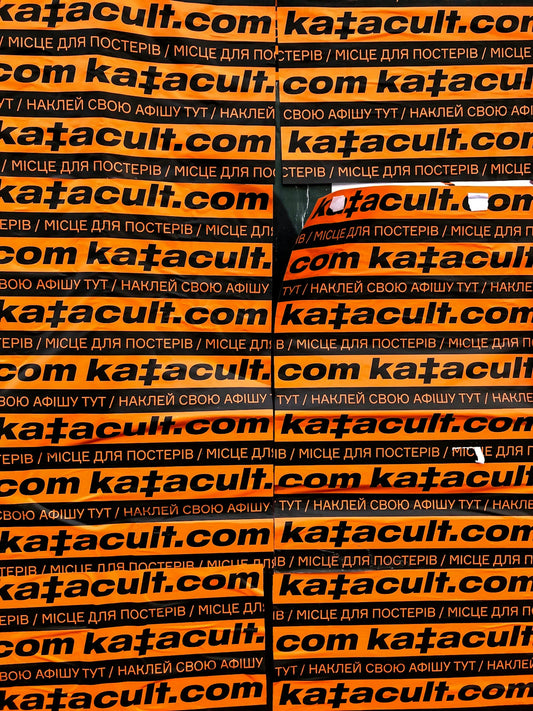 Orange posters with black text repeating 'katacult.com', covering a car headlight.