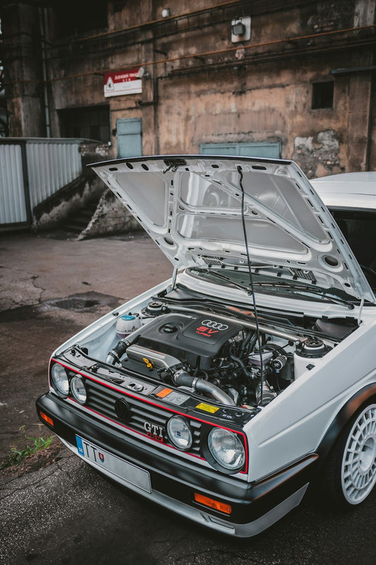 White Volkswagen GTI with hood open showing turbocharged Audi engine, urban background