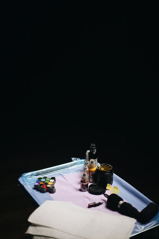 Tattoo supplies including ink bottles, machine, and ointment on a tray against black background.