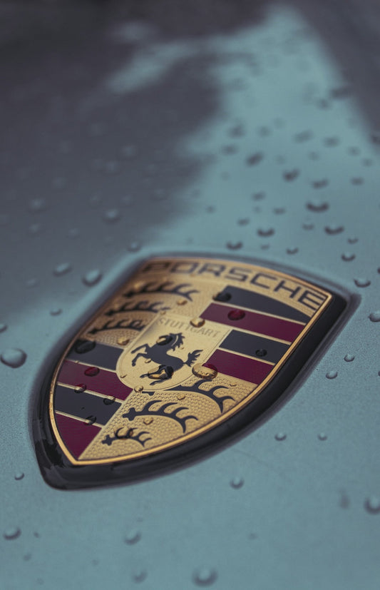 Close-up of Porsche car badge on hood with water droplets, luxury sports car emblem