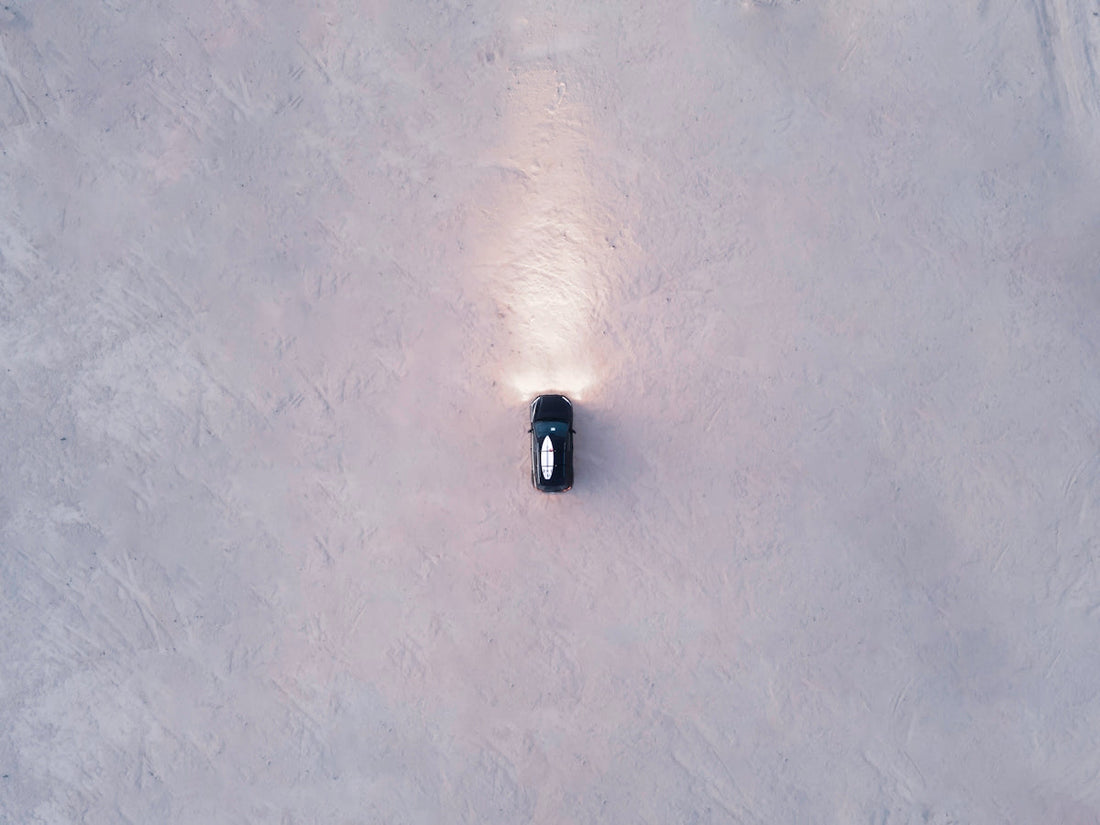 Aerial view of a black supercar driving on a sandy surface with headlights on