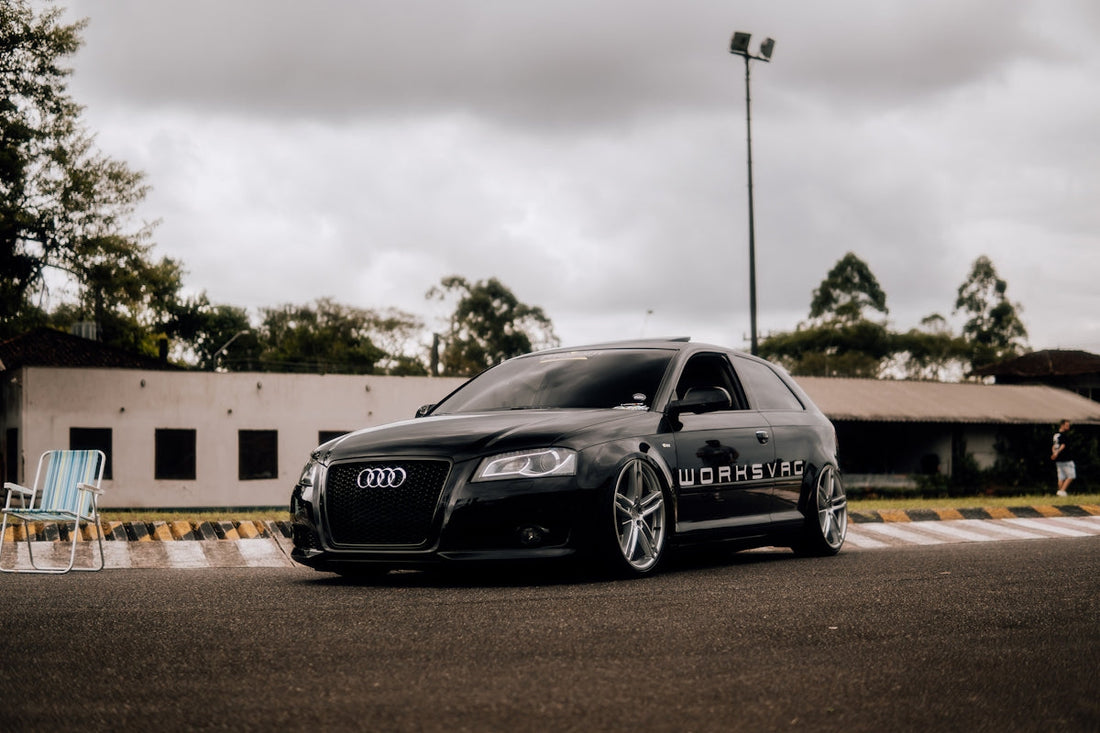 Black Audi hatchback with large alloy wheels on a street, overcast sky, tuned car exterior