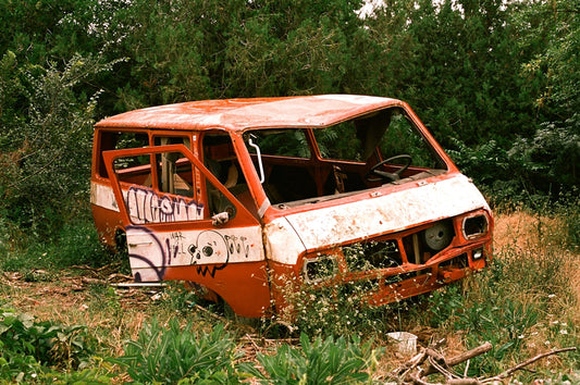 Abandoned, graffiti-covered van with missing parts in overgrown grass and trees