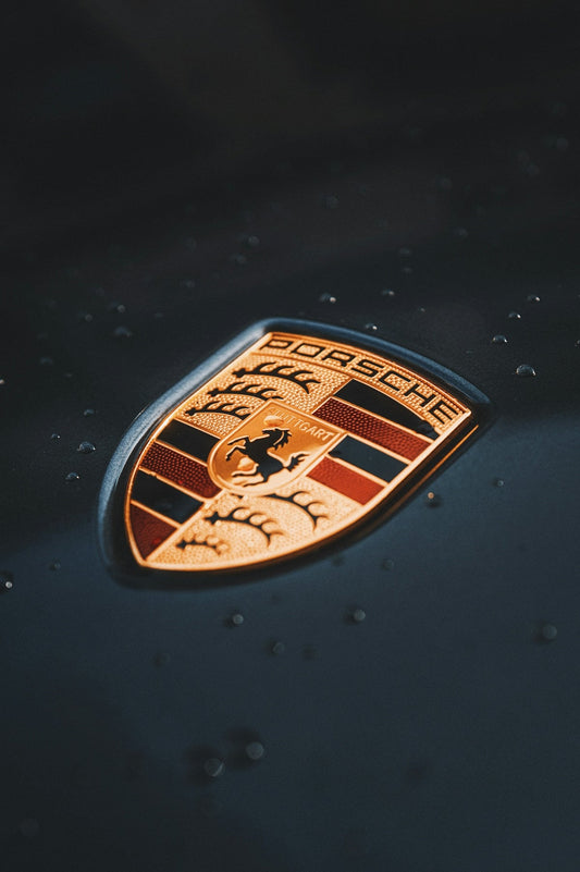 Close-up of Porsche emblem on dark car hood with water droplets, luxury sports car badge