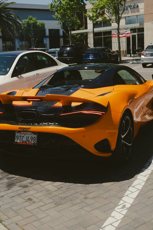 Orange McLaren supercar parked outdoors, rear view, featuring black accents and rear spoiler.