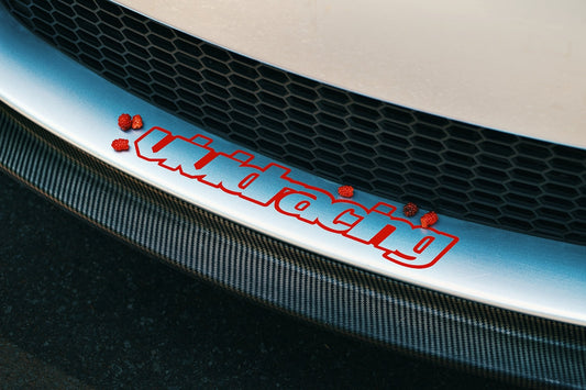 Close-up of car front bumper with 'vividracing' decal, carbon fiber splitter, and raspberries