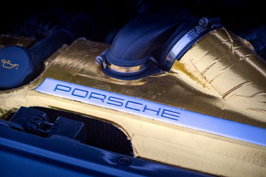 Close-up of Porsche engine cover with gold heat shield and metal branding emblem