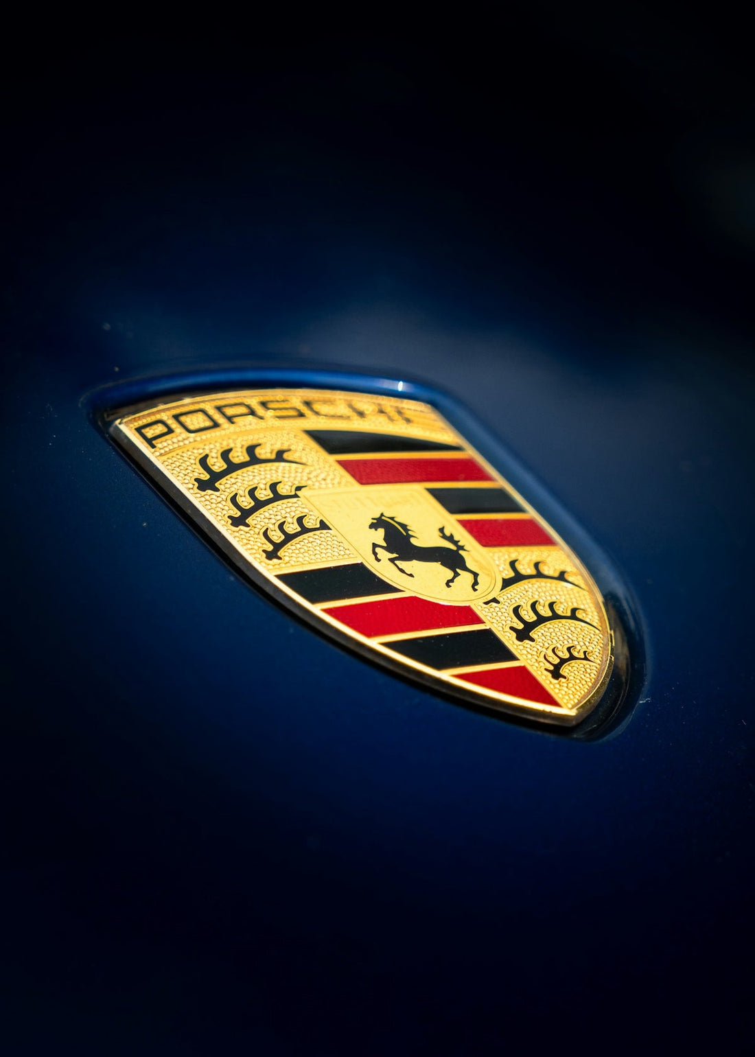 Close-up of a Porsche car emblem on a glossy dark blue hood, luxury supercar detail