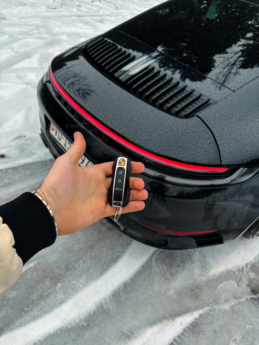 Hand holding Porsche key fob near black sports car on snowy ground